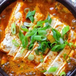 A close-up of a spicy doenjang jjigae topped with chopped green onions. The tofu is sliced and immersed in a rich, red broth, garnished with green onions, adding color and texture. Bubbles in the stew indicate it is hot and freshly cooked.